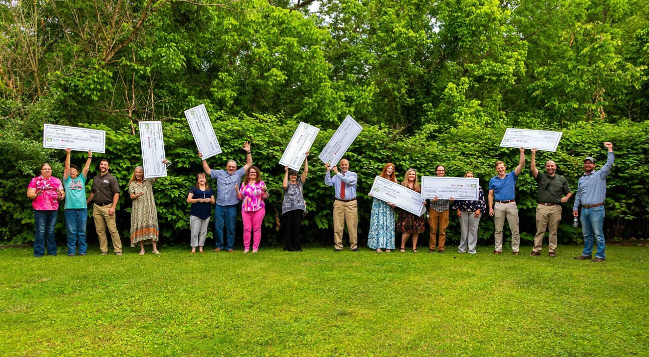 Award recipients stand on vivid green grass and in front of green trees, holding large checks.