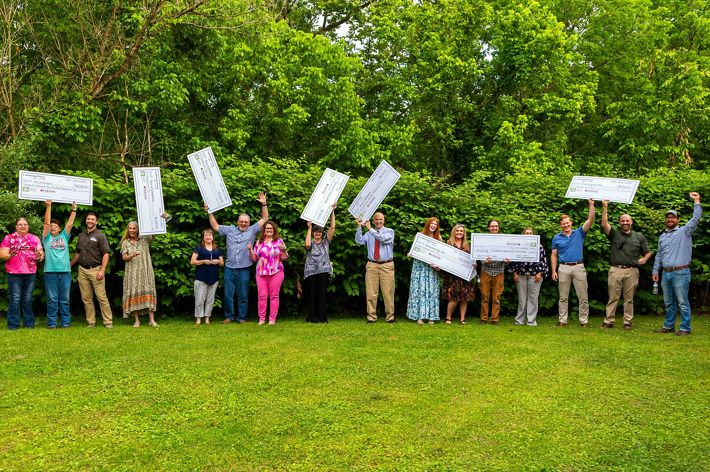 A group of people people pose together outdoors backed by tall trees. Many of the people hold aloft large, novelty checks given out during a grant award ceremony.