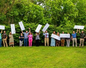 Award recipients stand on vivid green grass and in front of green trees, holding large checks.