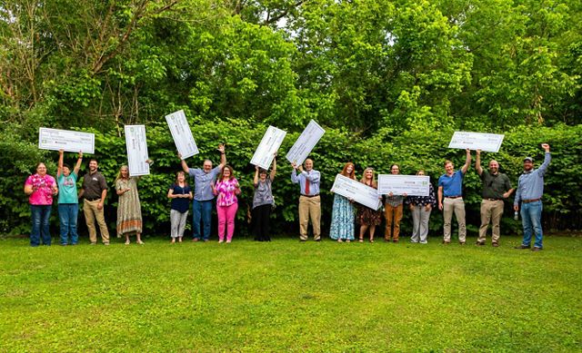 A group of people people pose together outdoors in a grassy era backed by tall trees. Many of the people hold aloft large, novelty checks given out during a grant award ceremony.