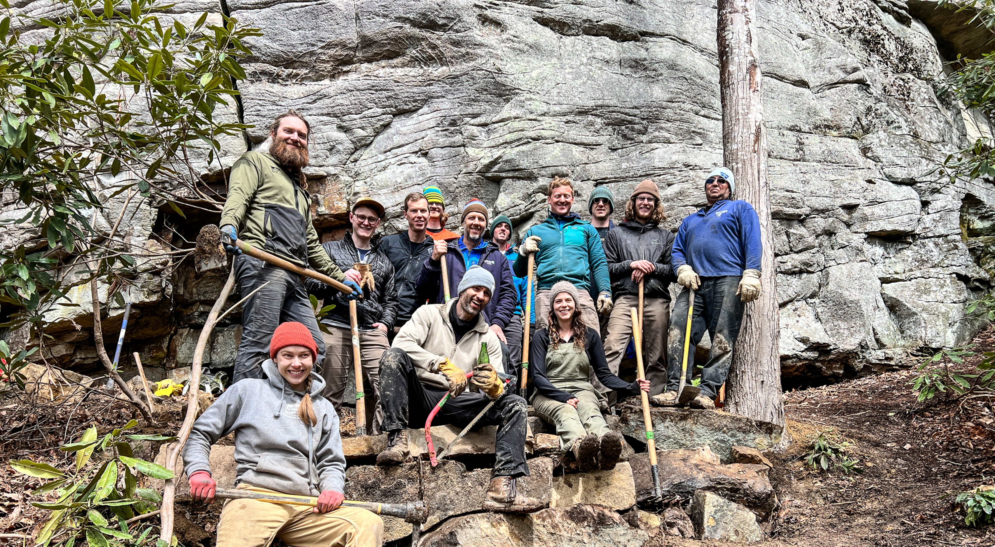 A large group of people pose together in front of a rock wall. They hold loppers and other hand tools for clearing brush and small vegetation.