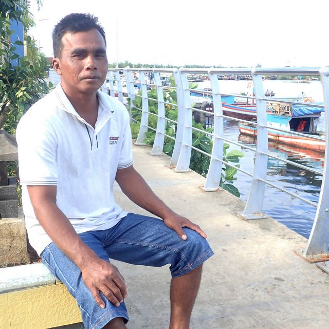 An Indonesian man sits on a dock with boats behind him.