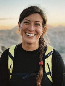 A woman with hiking gear smiling at a camera outdoors.