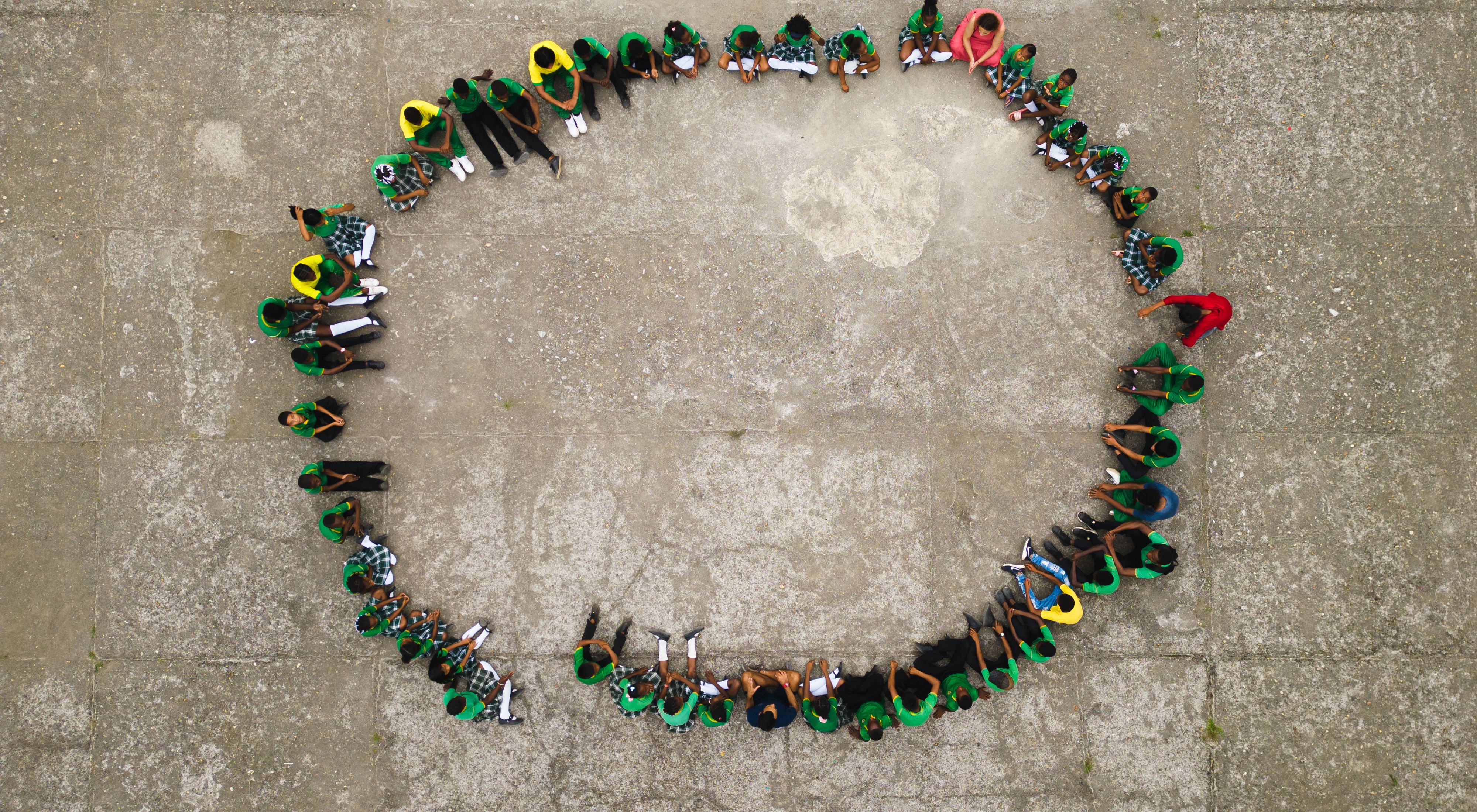 Aerial view looking down at a large group of teachers and students sitting in a large circle on the ground.