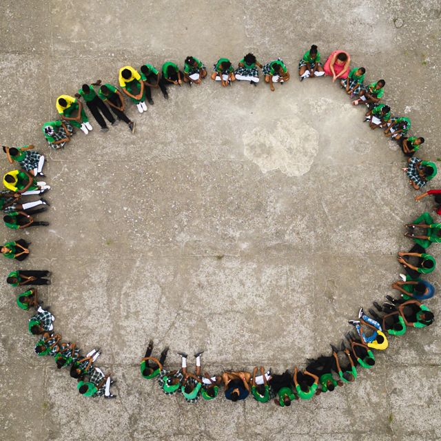 Aerial view looking down at a large group of teachers and students sitting in a large circle on the ground.