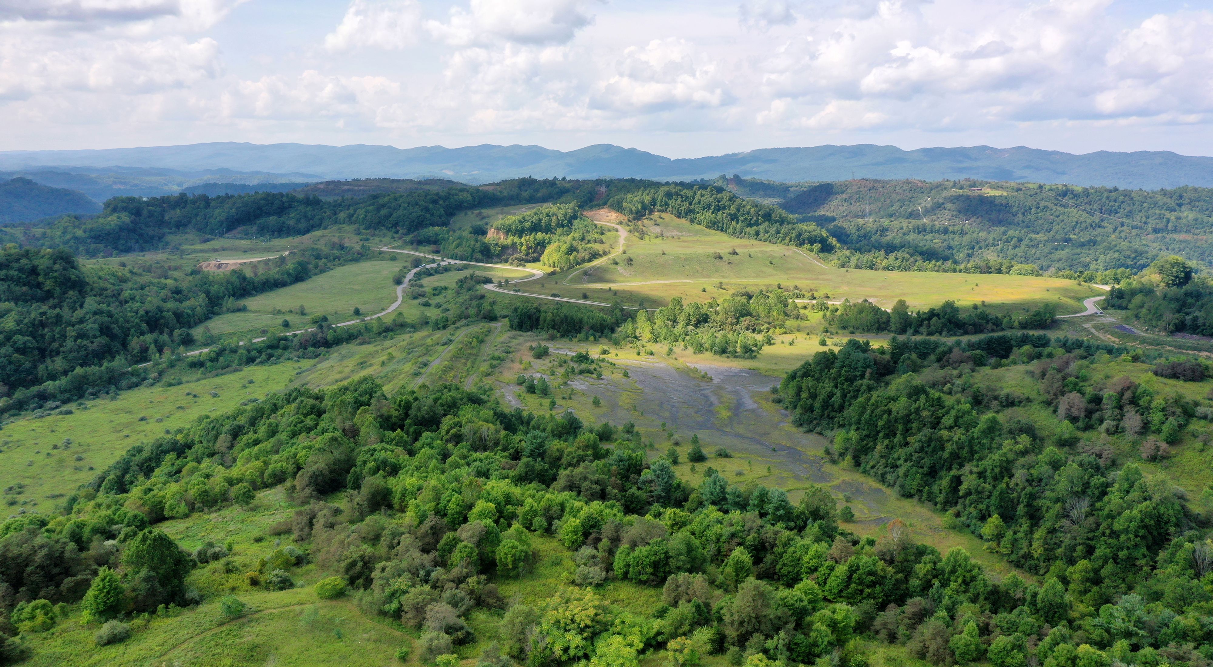 Aerial view of former mine lands in SW Virginia. A dark, bare earth scar lays in the center of an open field that is being reclaimed by grass. The patch is circled by trees and ringed by paved roads. 