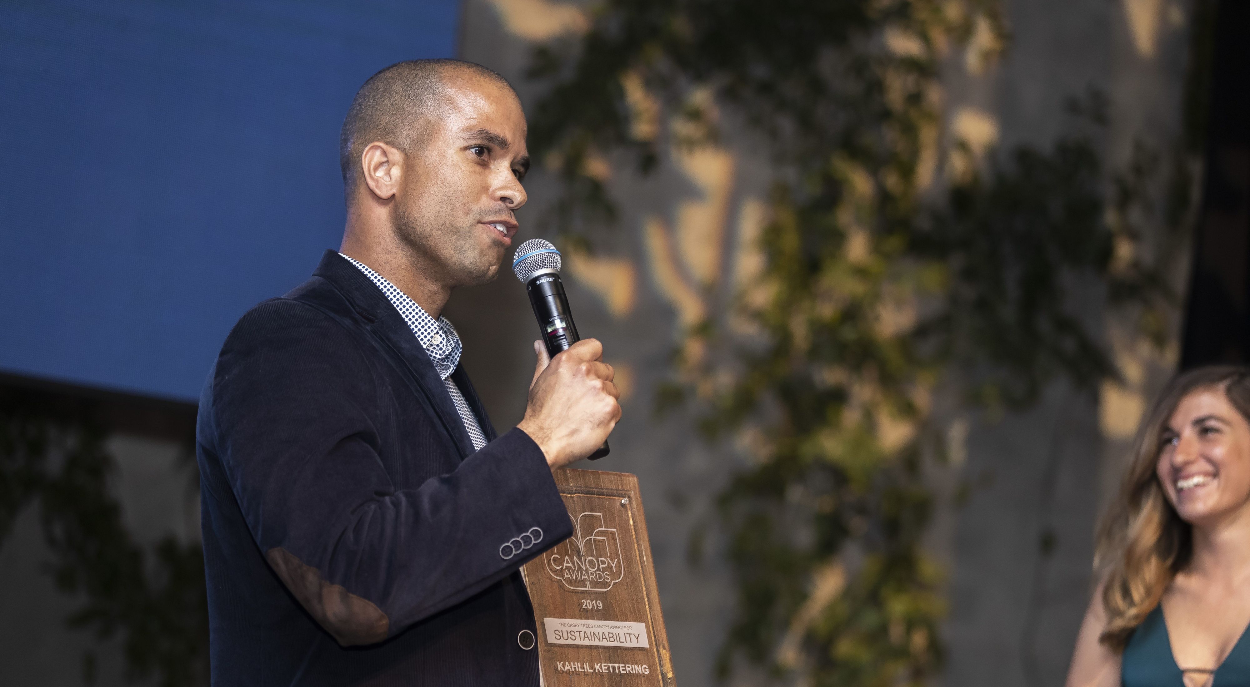  A person speaks into a microphone that they are holding and an award in the other hand.