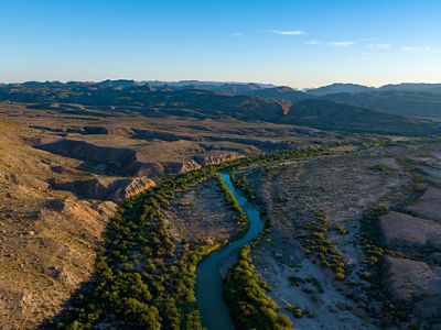 A blue river lined with brush winds through arid cliffs.