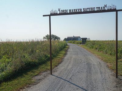 A tall metal entrance sign leads to a gravel trail through a field of native grasses.