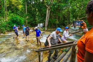 A woman takes a photo of people walking out of the water in the jungle.