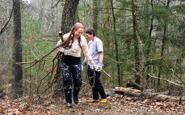 Two people walk in the forest carrying cut branches and vegeation.