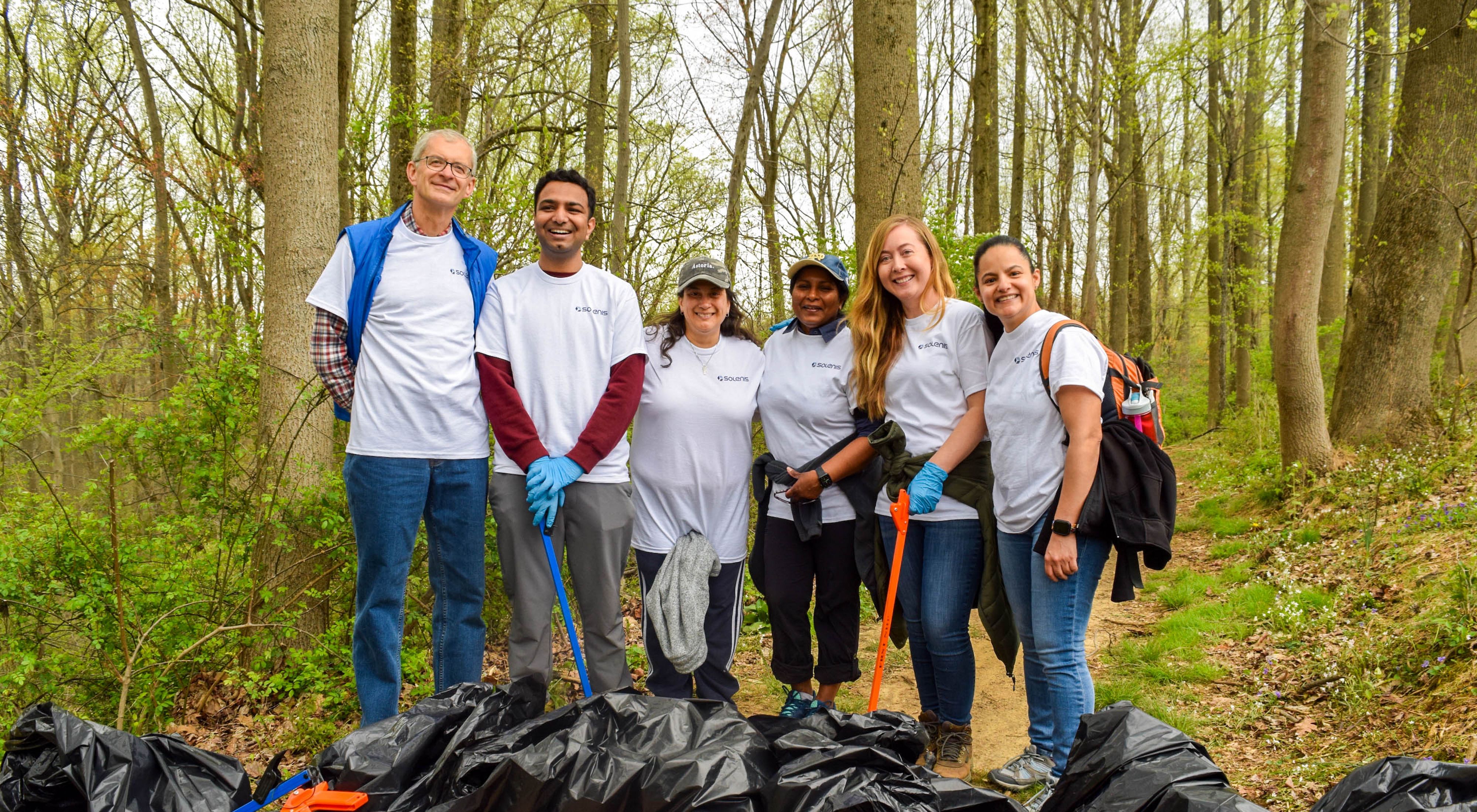 Six people pose together during a stream cleanup event. They are holding large black plastic trash bags and carrying long orange grabbers.