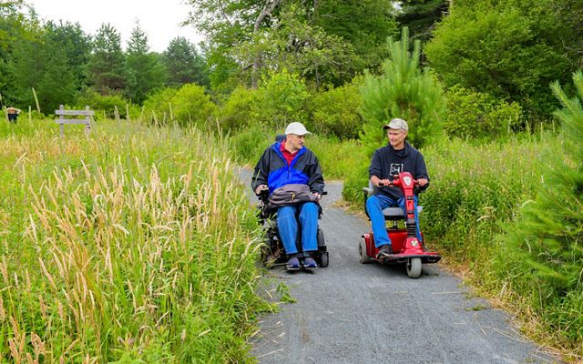 Two people using wheel chairs talk to each other as they explore a nature trail.