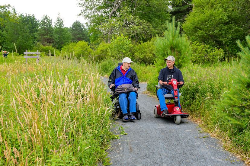Two people using wheel chairs talk to each other as they explore a nature trail.