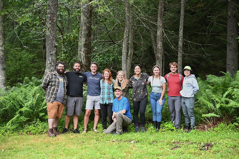 A group of 10 people pose together arm in arm at the edge of a forest on Warm Springs Mountain.