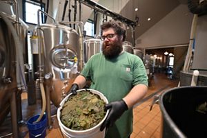 A man stands in a brewery next to a large open tank. He holds a large bucket filled with green leaves.