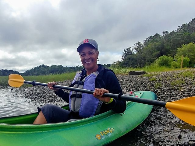 Dale Threatt-Taylor sostiene un remo dentro de un kayak, en una orilla frente a un paisaje boscoso.
