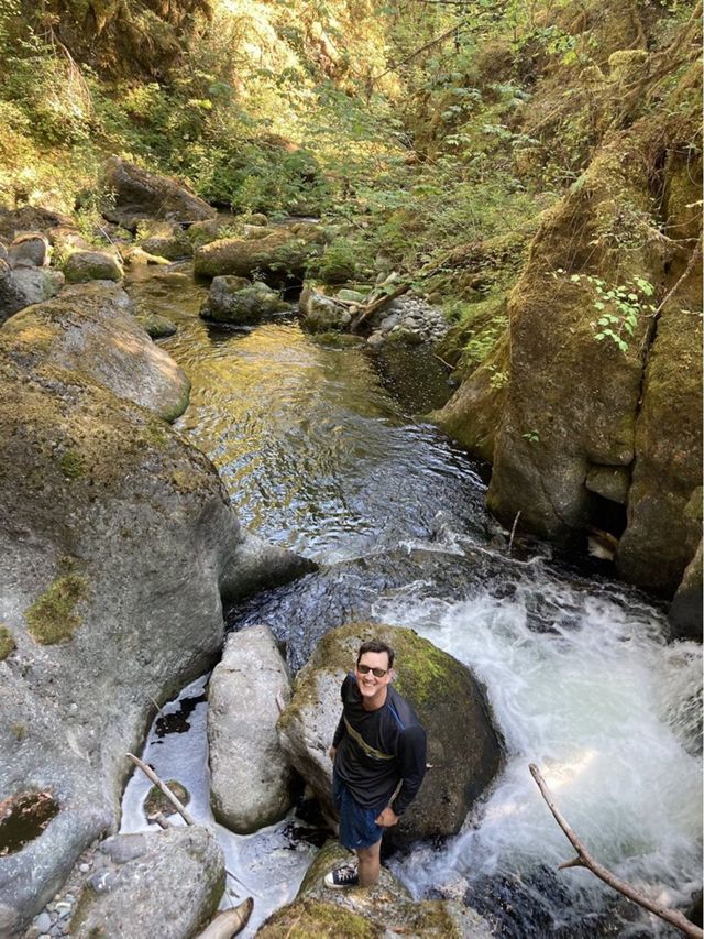 A man stands on a large rock in the middle of a forest creek. The water foams white as it rushes through the narrow channel.