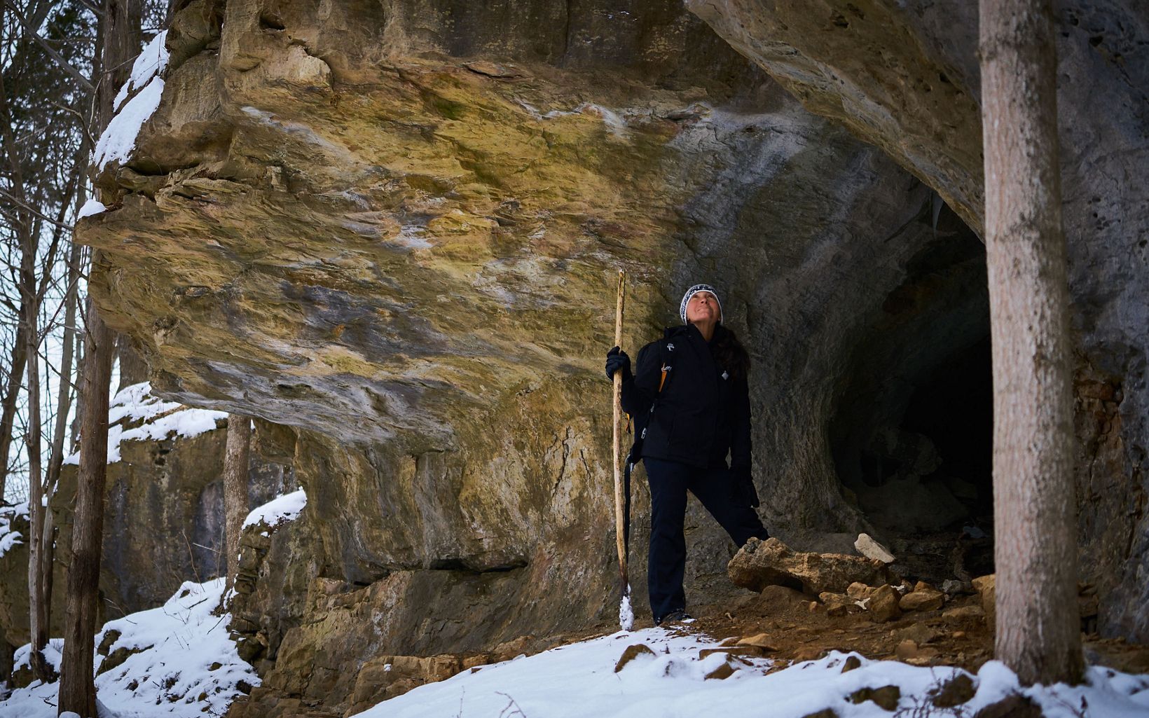 A hiker pauses under a rock outcrop in the snow. 