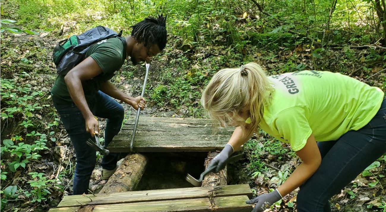 Two people working on a footbridge in the woods.
