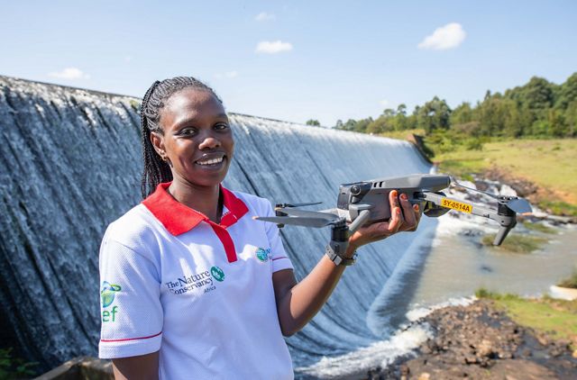 A woman holding a drone stands in front of a dam. 