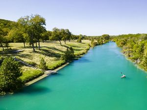 A kayaker on a bright turquoise Blanco River running between one grassy shore and one that is lined with trees.