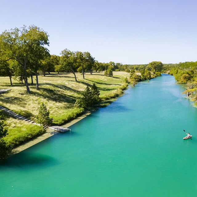 A kayaker paddles on a turquoise river lined with trees and green grass.