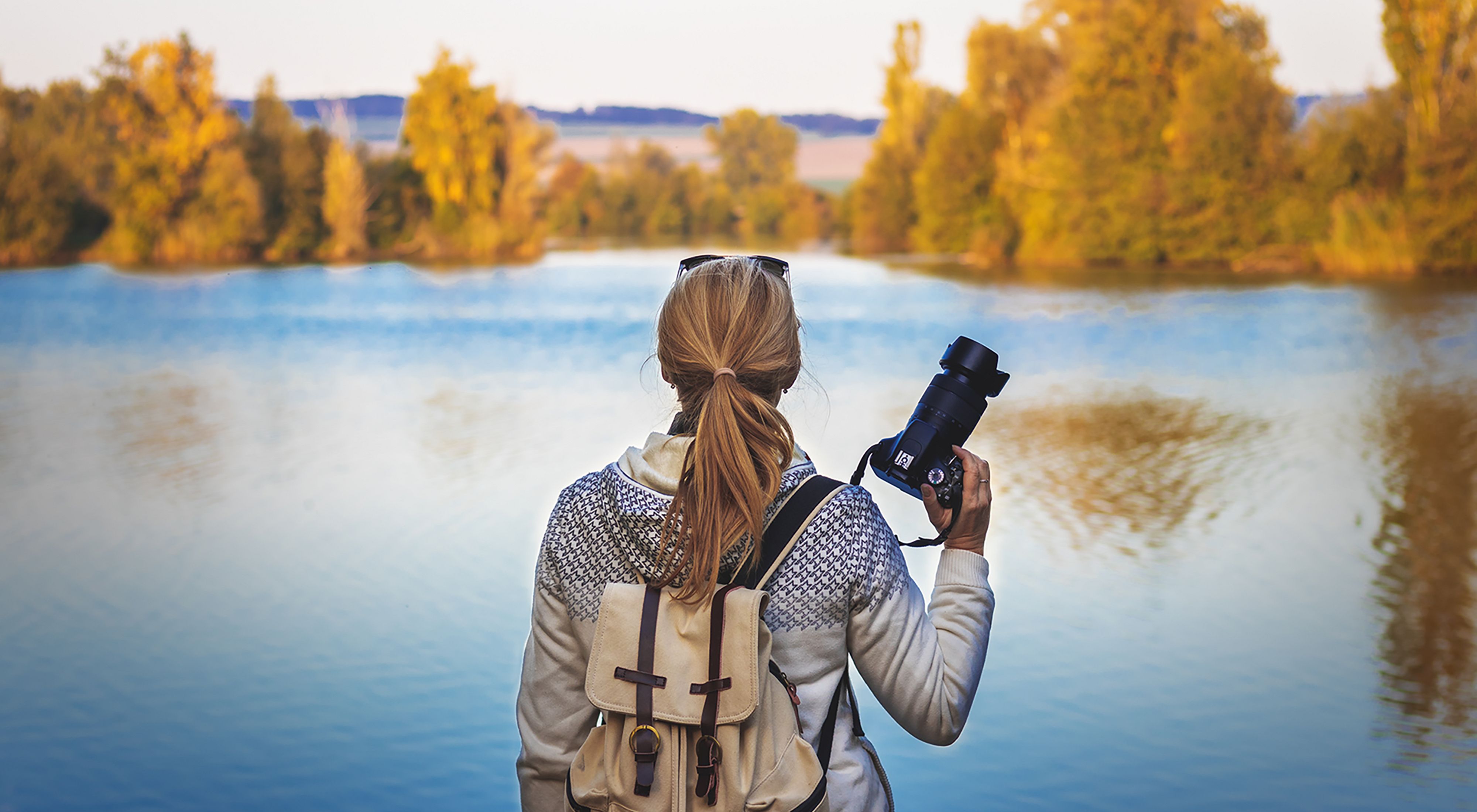 A person who is holding a camera looks out on a still lake that is surrounded by green trees. 