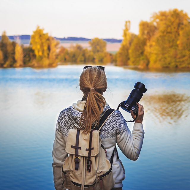 A person who is holding a camera looks out on a still lake that is surrounded by green trees. 