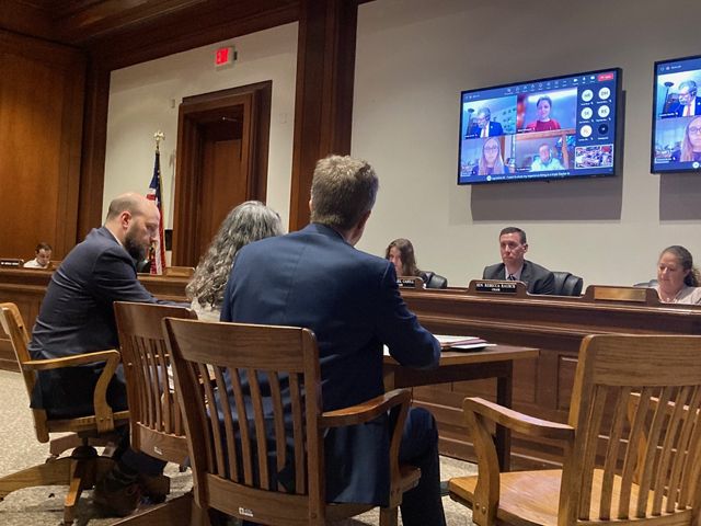 Three people sit at a table testifying for legislation to members of a Masscahusetts House Committee, sitting at al ong table. The screen behind them shows a Teams call where people joined virtually.
