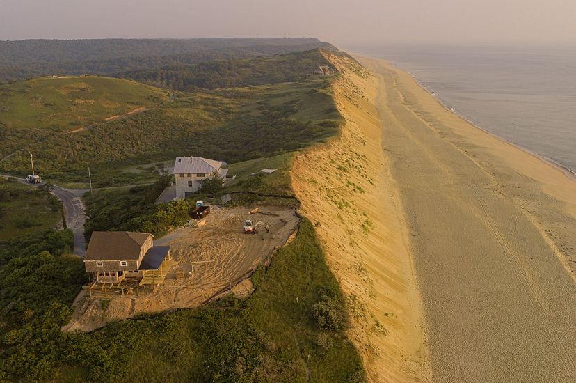 a steep sand bluff is seen from above with two houses near the edge. One house sits farther back from the edge with construction equipment nearby