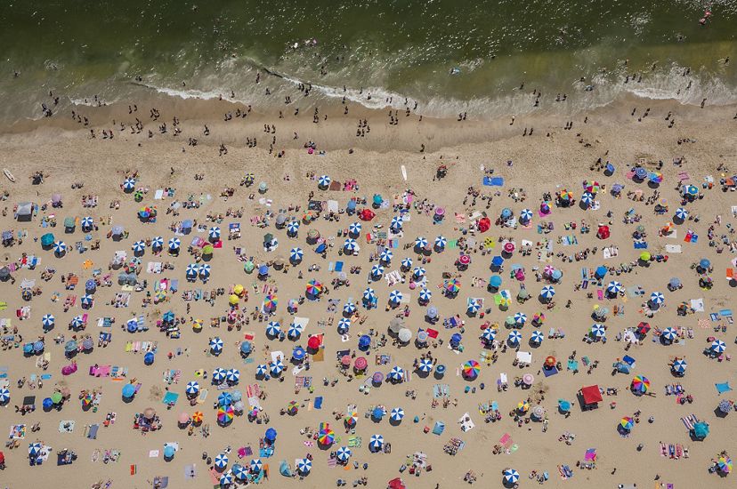 Seen from above: dozens of beach umbrellas and towels dots a beach with swimmers standing on the edge of the water
