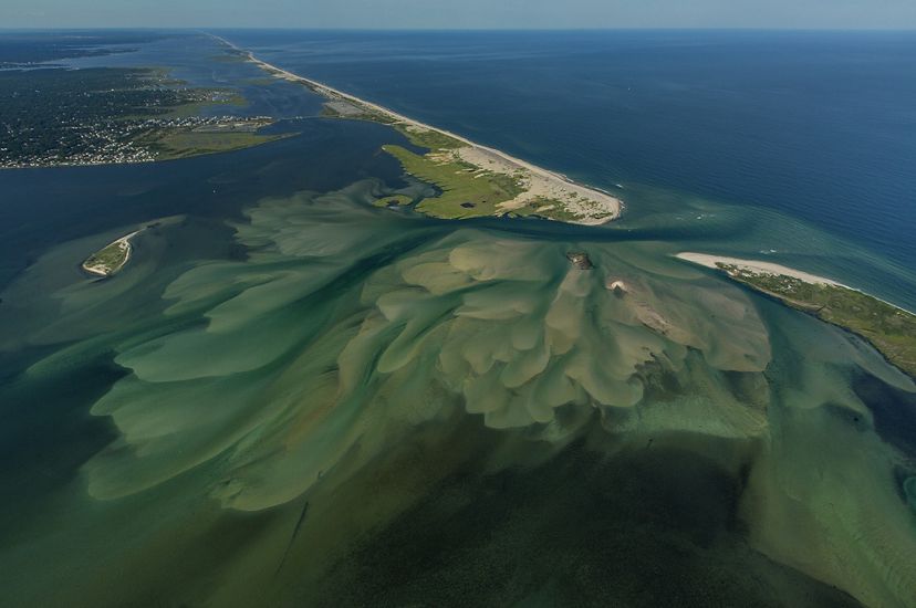 seen from above: a narrow stretch of land surrounded by blue ocean water on both sides is shown with a cut in the middle of the land where the water has pushed through, mud/sand visible on the water