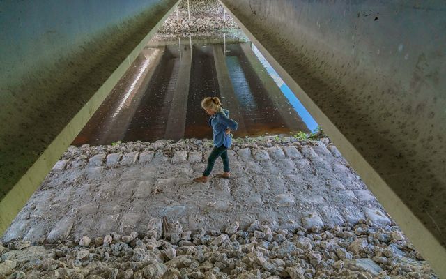 woman stands under highway bridge on a rocky path near a water-filled canal in Florida