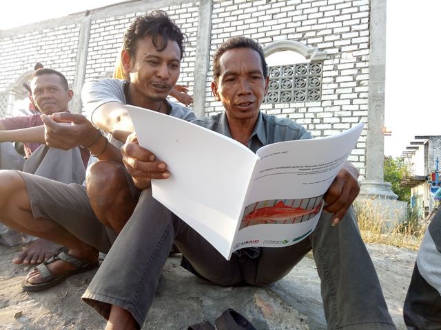 Three men sit on the ground and look at a book about fish.