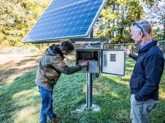 Two men stand next to a small solar panel that powers a suburban smart storm pond. 