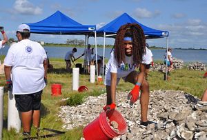 A woman holds a red bucket to scoop up oysters from a larger pile.