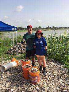 A man and woman stand in a pile of oyster shells with orange buckets also filled with oyster shells.