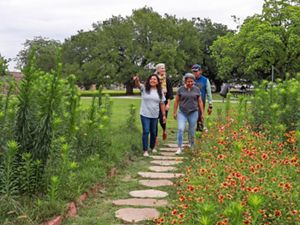 Three women and one man walk on a path with flowers in a green park.