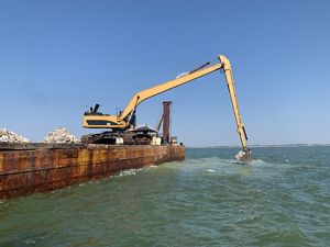 An excavator sits on a barge in the ocean, dumpig limestone boulders in a bay.