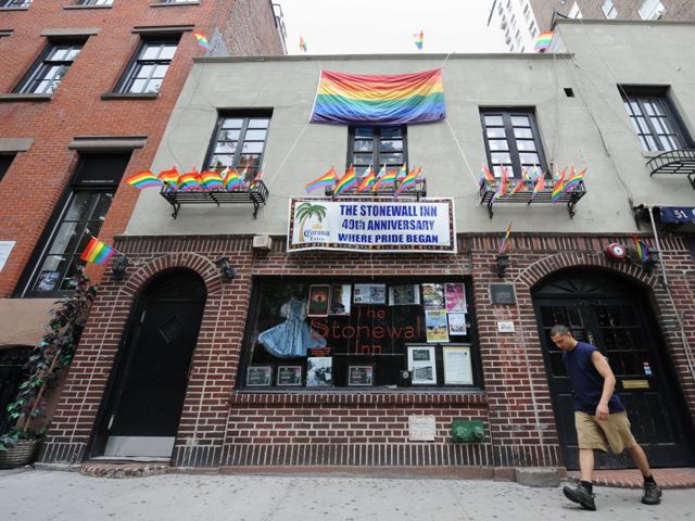A man walks passed the urban business storefront of NY's Stonewall Inn. Two arched doorways flank a wide picture window. The tall windows above the entrance are overhung with a rainbow Pride banner.