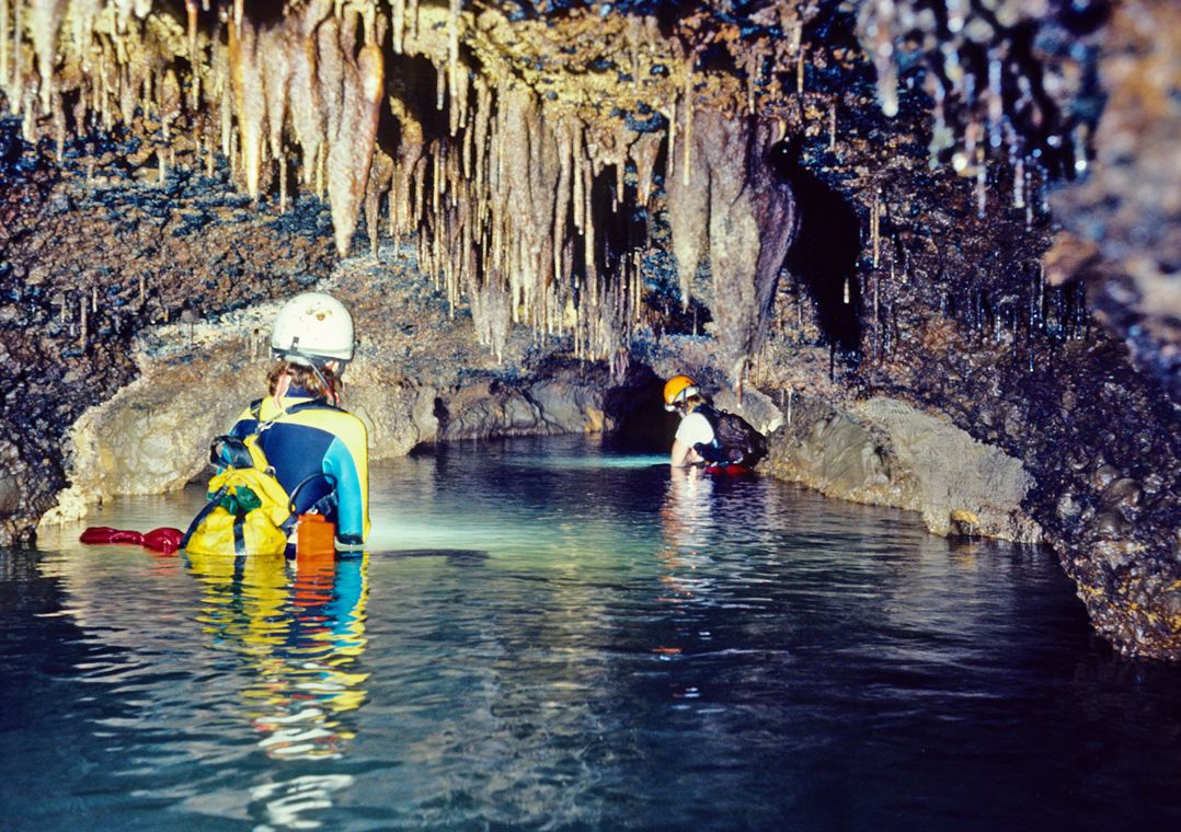 Two people in helmets wade through water in a cave.