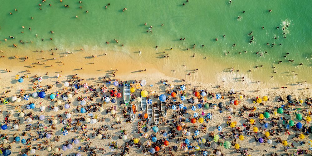 Decenas de personas, kayaks, sombrillas y toallas se agolpan en la playa. Algunas personas nadan en las olas bajas.
