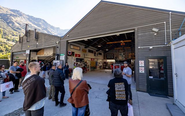Several people stand outside a boathouse with its doors open as one person talks to the group.