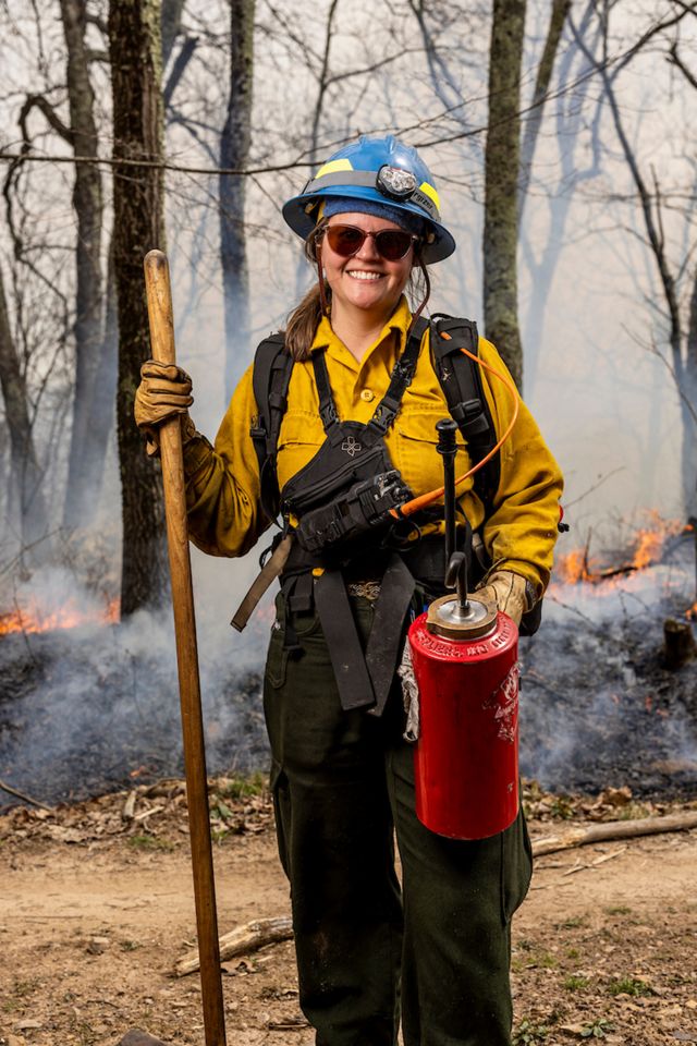 A smiling woman poses in front of a prescribed burn in fire protective clothing.