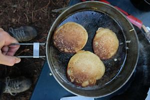 Three flapjacks in a skillet being cooked over a campfire. 