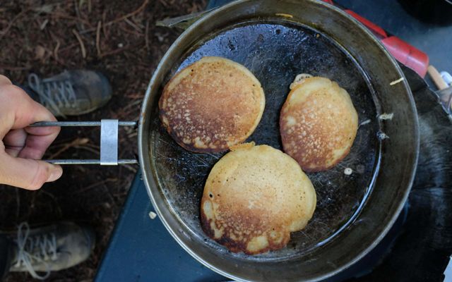 Three flapjacks in a skillet being cooked over a campfire. 