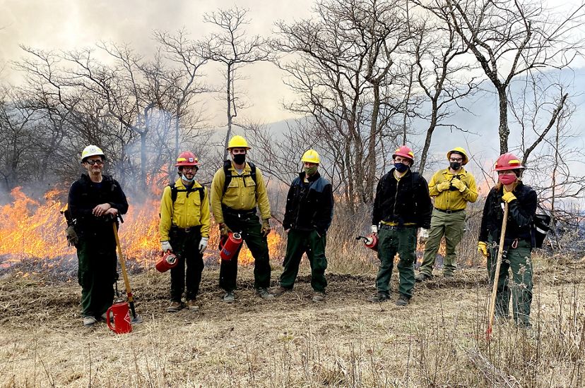 Seven people pose together in a line during a controlled burn. They are wearing yellow fire gear and some hold red drip torch canisters. Fire burns through the tall grass behind them.