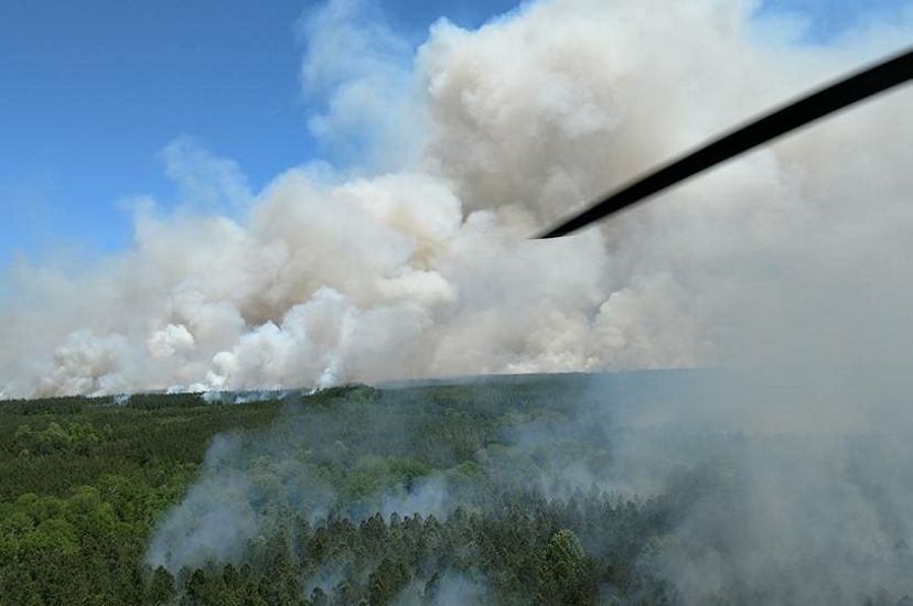 Aerial view of a controlled burn. Thick white smoke billows above a wide expanse of forest. A helicopter rotor blade slices through the air.