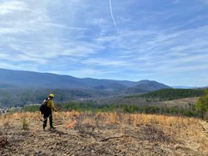 A man wearing yellow fire gear stands in an open field next to a small fire that is burning itself out. Tall mountain ridges line the horizon in the background under a bright blue sky.